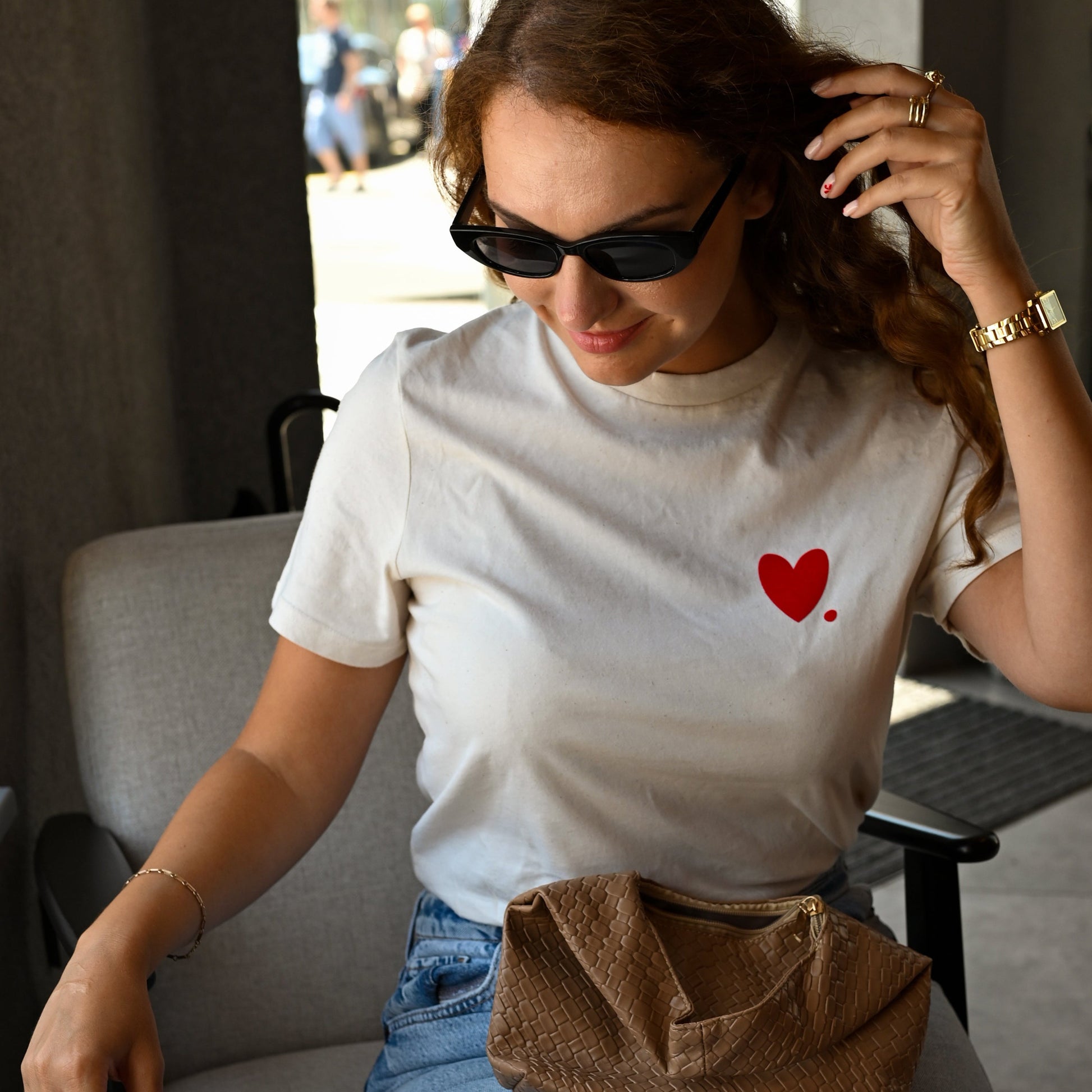 Woman sitting on a chair wearing a white t-shirt with a red heart design, blue jeans, and sunglasses.